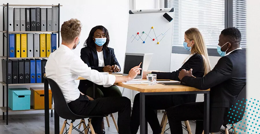 imagem de 4 pessoas ao redor de uma mesa todas usando mascara e conversando dentro de uma ambiente de trabalho 