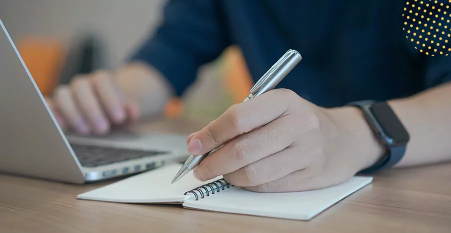 uma pessoa mexendo no notebook e escrevendo num papel sob a mesa