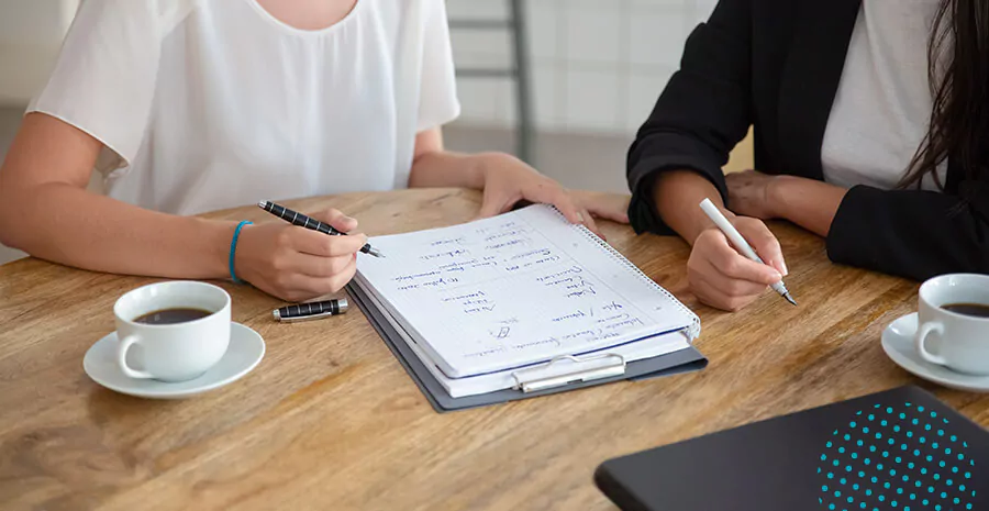 imagem de uma mesa com xícara de café, duas pessoas segurando uma caneta e um caderno ao centro