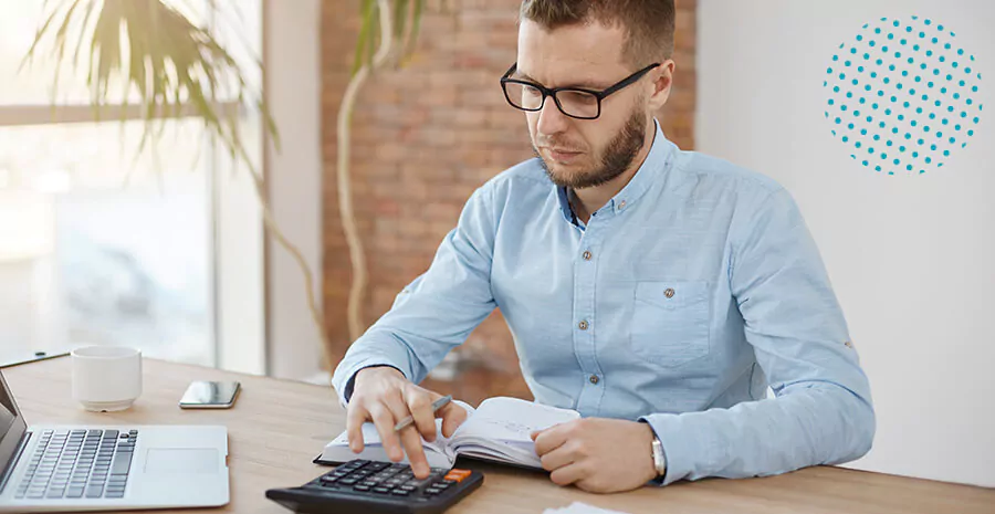 imagem de um homem mexendo na calculadora segurando uma caneta próximo a um caderno sob uma mesa com notebook e uma caneca 