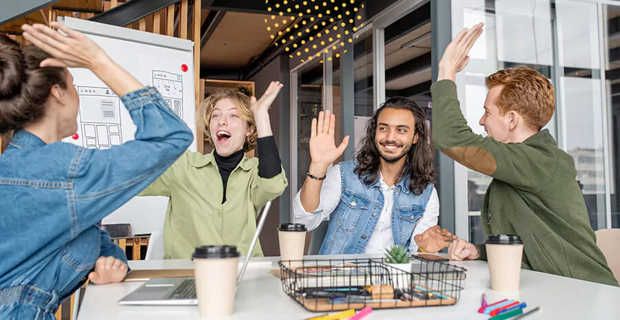 imagem de quatro pessoas comemorando em volta de uma mesa de trabalho com um quadro branco atrás