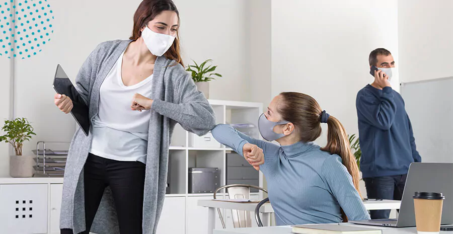 imagem de duas mulheres de máscaras no ambiente de trabalho se cumprimentando pelos cotovelos 