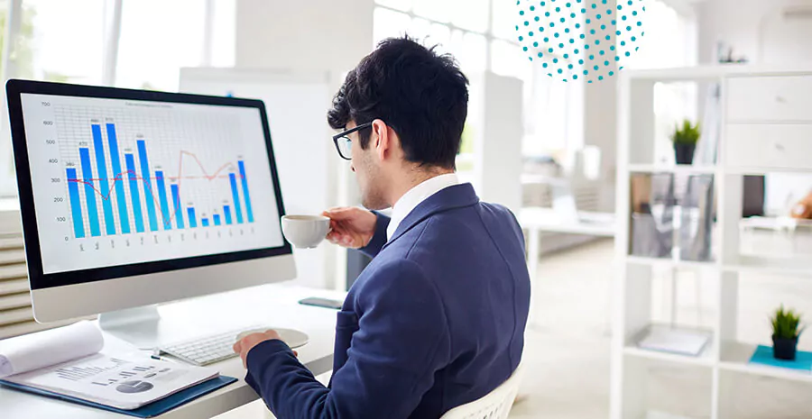 imagem de um homem sentado no ambiente de trabalho tomando café, em frente a uma tela de computador mostrando gráficos