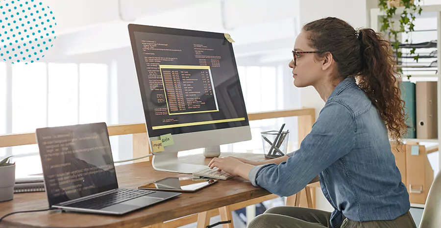 imagem de uma mulher cto de uma empresa sentada mexendo num computador com uma tela e um notebook ao lado com códigos 