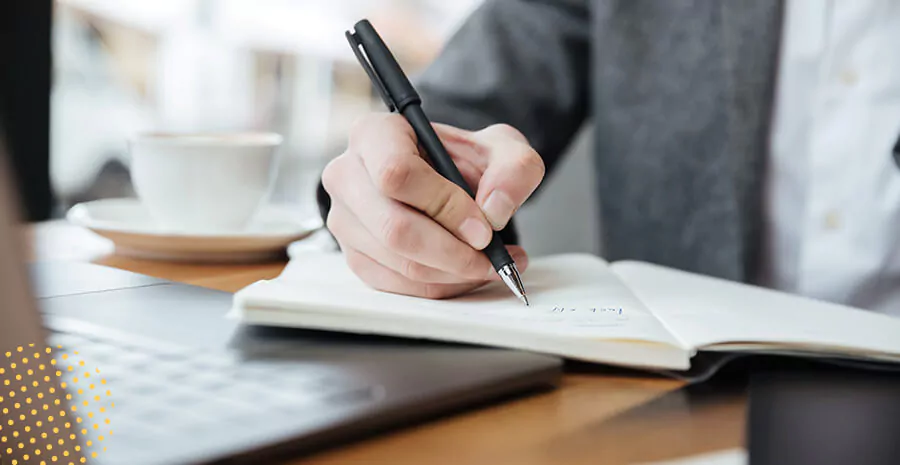 imagem de uma mesa com notebook e xícara de café desfocados e uma pessoa escrevendo em um caderno com uma caneta