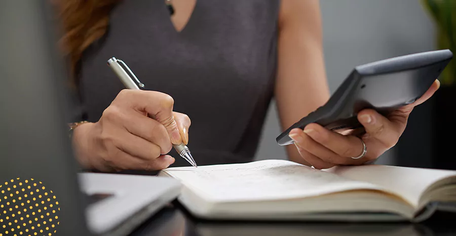 imagem de uma mulher segurando uma calculadora e anotando com uma caneta em um caderno