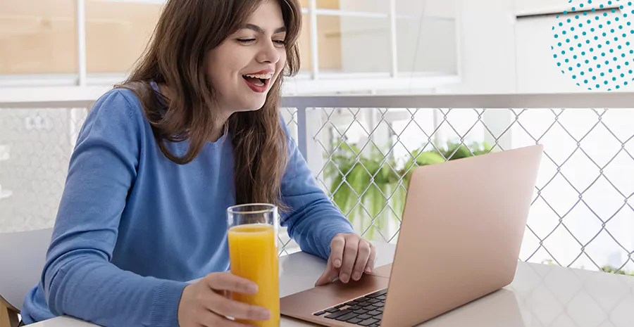 imagem de uma mulher sentada na frente do computador sorrindo e bebendo um suco