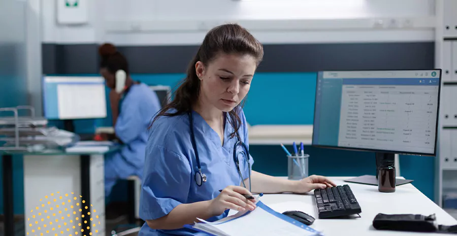 imagem de uma enfermeira sentada na frente de um computador olhando para uma folha de papel ao lado