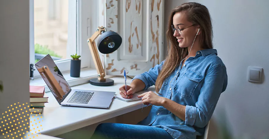 imagem de uma mulher sorrindo sentada na frente de um computador e escrevendo em um caderno