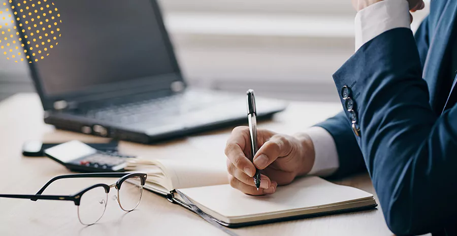 imagem de um homem sentado escrevendo em um caderno com um computador e um óculos ao lado na mesa