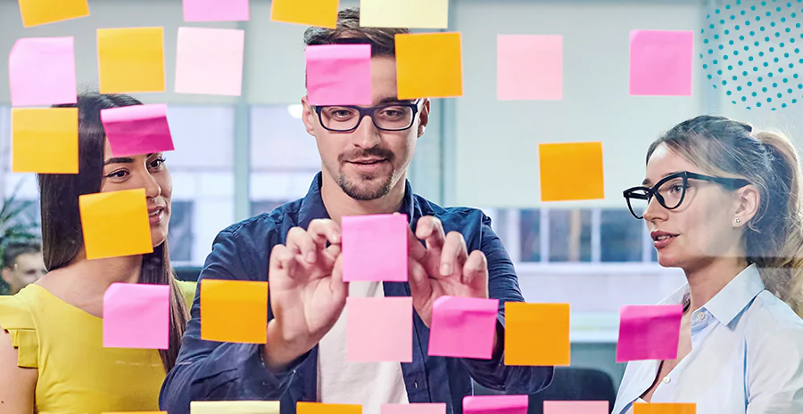 imagem de um homem e duas mulheres olhando para postits grudados em uma parede de vidro