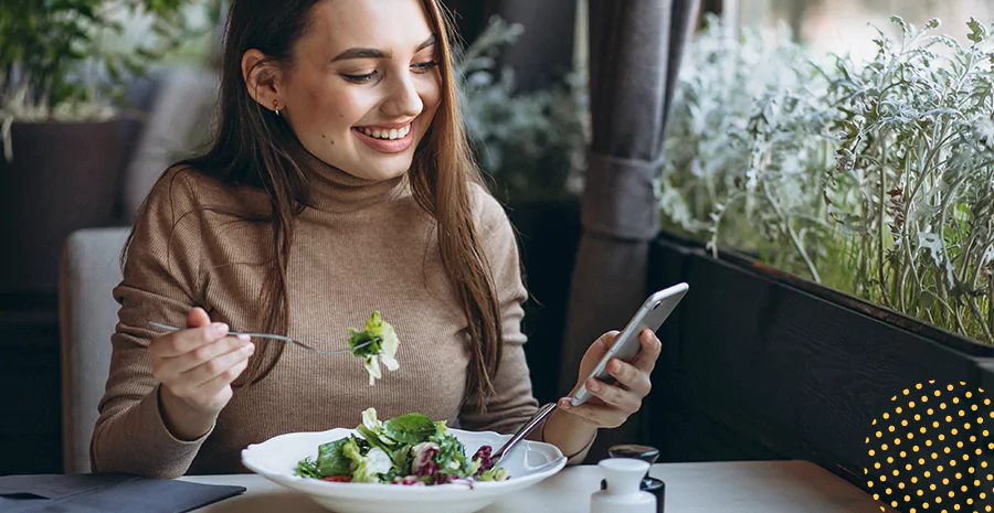 imagem de uma mulher sentada sorrindo comendo salada e segurando um celular