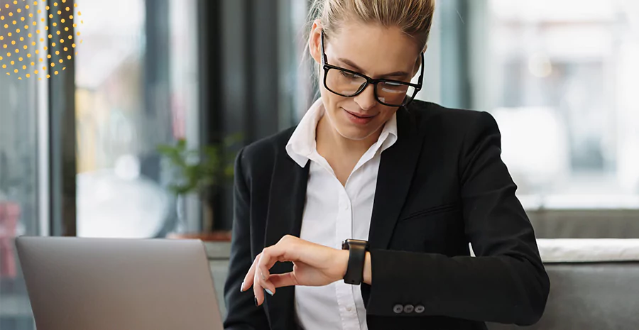 imagem de uma mulher sentada na frente de um computador olhando para seu relógio de pulso