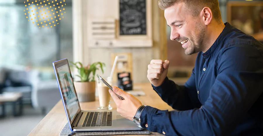 imagem de um homem sentado na frente de um computador sorrindo e segurando um celular