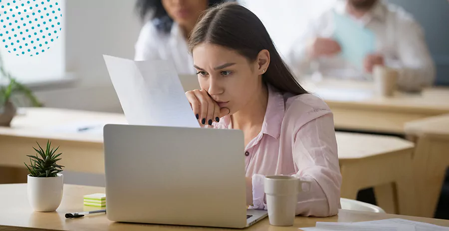 imagem de uma mulher séria sentada na frente de um computador e olhando para um papel