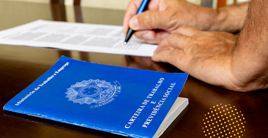 Um homem assinando um documento que está ao lado de uma carteira de trabalho