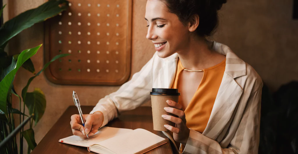 Mulher anotando em uma caderno enquanto toma café