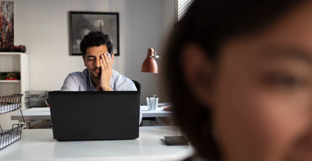 imagem mostra homem em frente ao computador e com uma das mãos na cabeça com expressão de descontentamento 