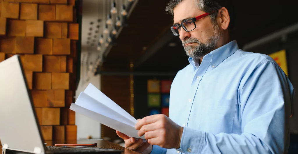 Um homem de óculos segurando um papel e olhando para um laptop