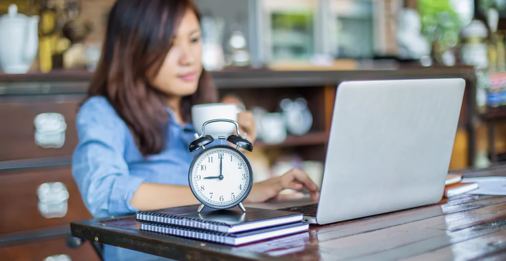 Um relógio e ao fundo Uma mulher sentada à mesa com um laptop