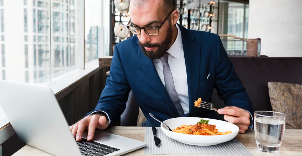 Um homem de terno comendo macarrão e usando um laptop