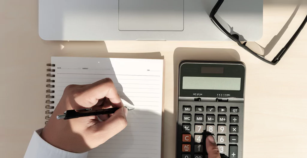 imagem mostra uma mão usando a calculadora e fazendo anotações em um caderno 