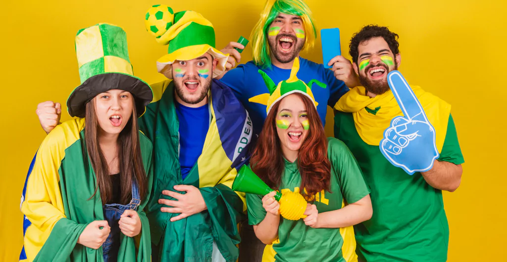 Grupo de cinco torcedores brasileiros animados, vestidos com roupas e acessórios nas cores da bandeira do Brasil, em fundo amarelo