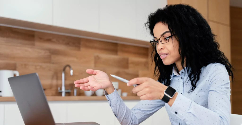 Mulher em cozinha moderna de escritório gestualizando durante videochamada no laptop, usando smartwatch