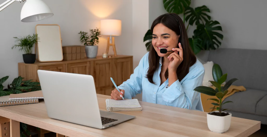 mulher sentada na frente de um notebook usando um fone de ouvido e fazendo anotações em um caderno 