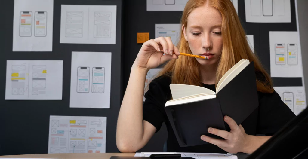 Mulher ruiva lendo caderno, com esboços de aplicativos móveis presos na parede ao fundo