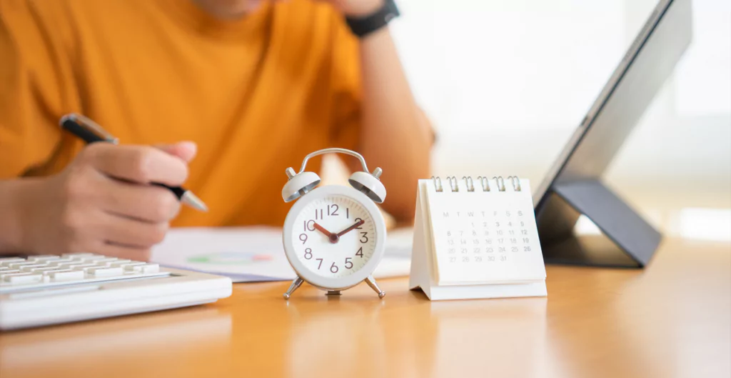 Relógio despertador e calendário sobre mesa, com pessoa escrevendo e usando laptop ao fundo.