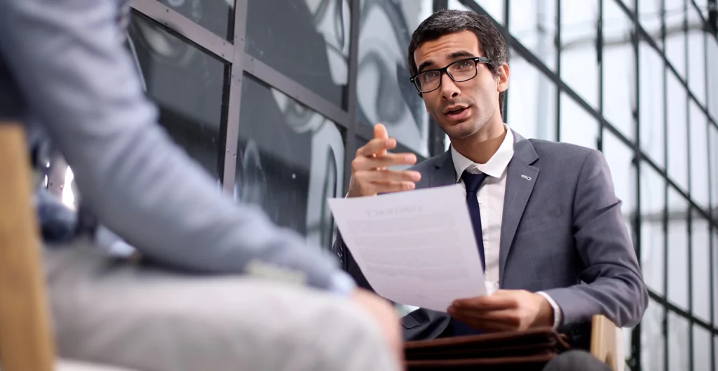 Homem de terno segurando documento e conversando durante entrevista de emprego.