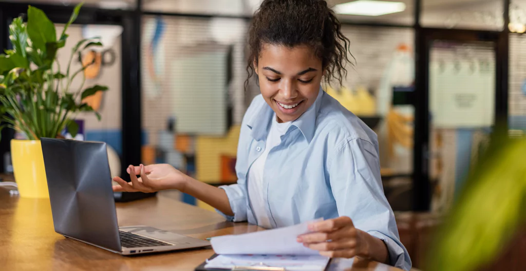 Mulher sorridente revisando documentos em frente ao laptop em escritório moderno