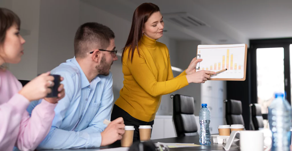 Mulher apresentando gráfico em reunião, enquanto dois colegas assistem e seguram copos de café