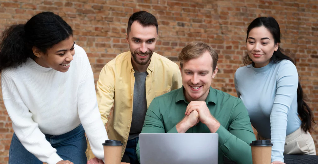 grupo de 4 pessoas, sendo 2 homens e 2 mulheres, sorridentes em frente de um notebook 