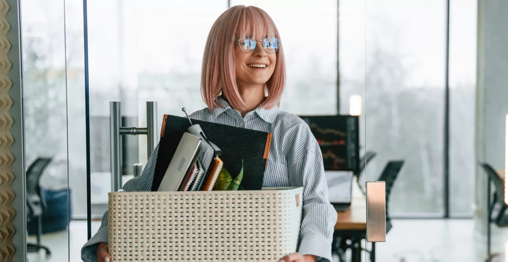 mulher sorridente carregando uma caixa com objetos de escritóro 