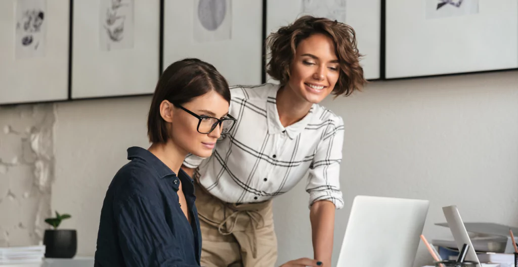 Duas mulheres colaborando em laptop, sorrindo e discutindo trabalho em escritório moderno