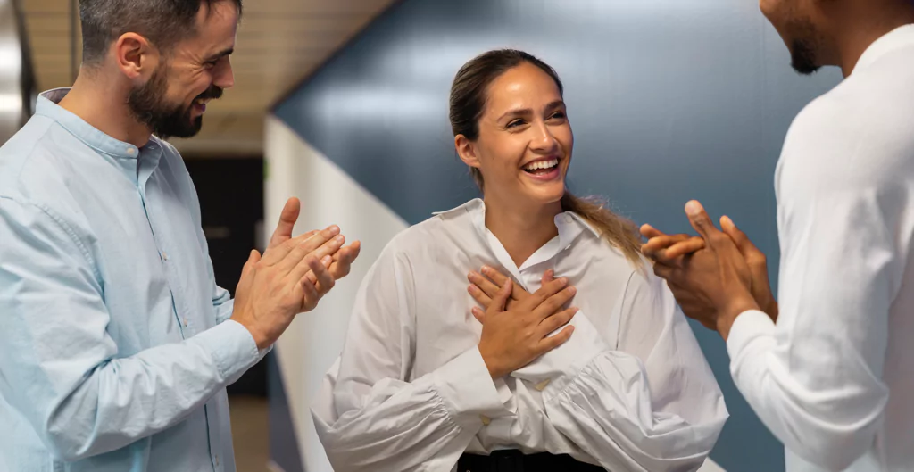 Mulher sorrindo e sendo aplaudida por dois colegas em corredor