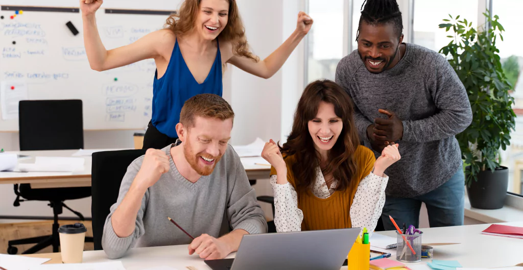 Equipe de quatro pessoas comemorando sucesso diante de laptop em escritório