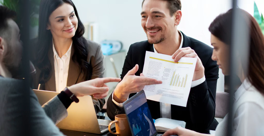 imagem foca em homem segurando um papel com informações em uma reunião com mais 3 pessoas 