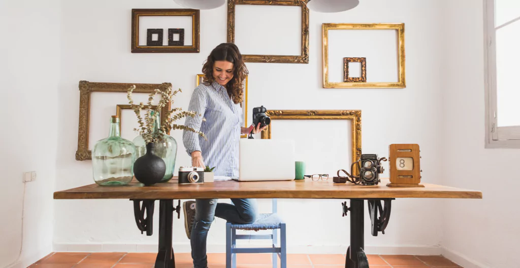 mulher em pé segurando uma câmera e de frente a uma mesa com um notebook e outras câmeras 