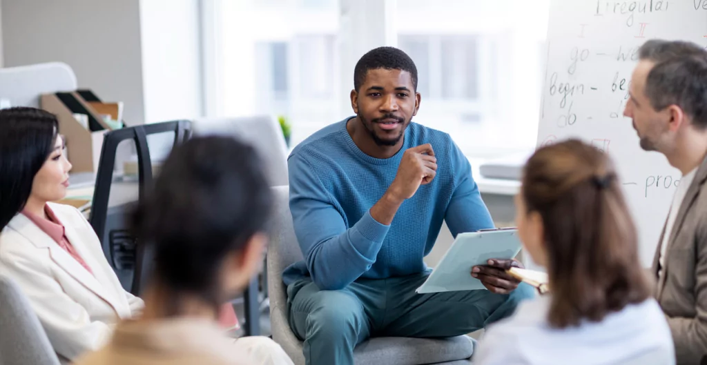 Homem fazendo apresentação em grupo, com quatro pessoas ao redor, em um ambiente de trabalho colaborativo.