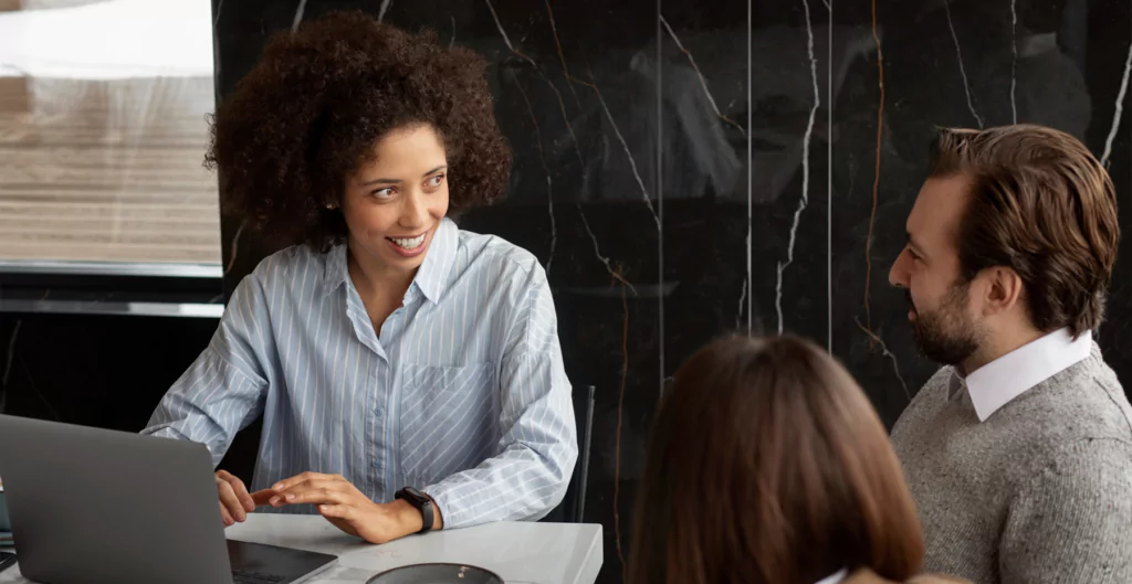 Mulher sorridente discutindo com dois colegas, usando laptop em uma mesa de reunião.