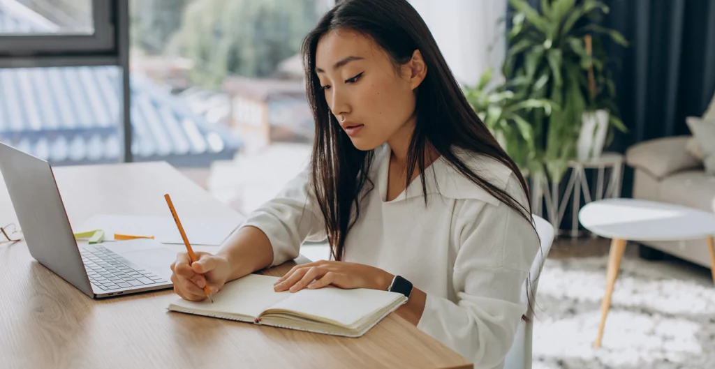 mulher sentada em frente a um notebook fazendo notações em um caderno 