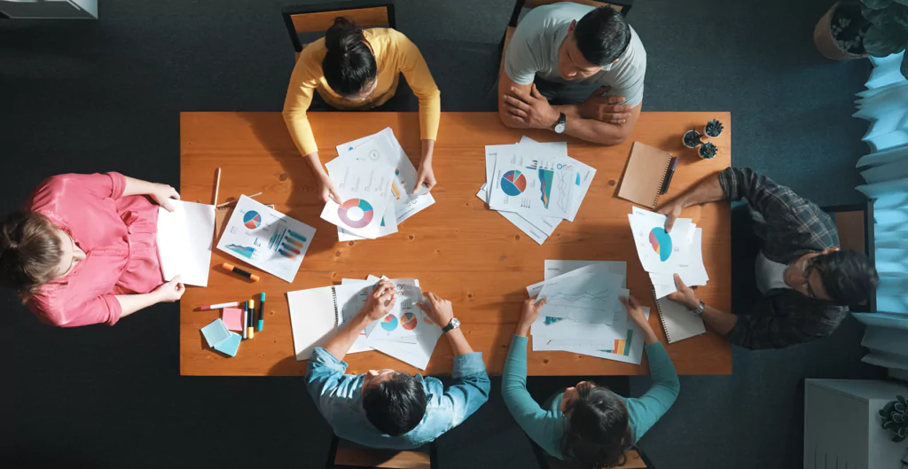 Grupo de pessoas analisando gráficos e documentos em uma mesa de reunião, visto de cima.