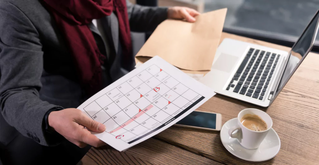 Pessoa em um escritório segurando um calendário impresso, com laptop e café ao lado.