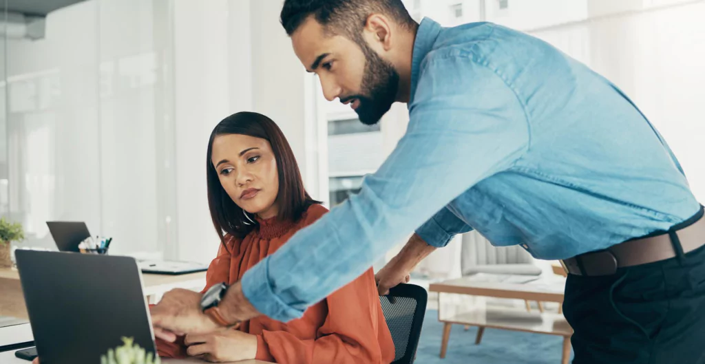 Mulher sentada de frente para um notebook conversando com um homem em pé ao seu lado 