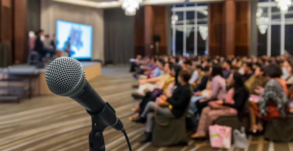 imagem com fico em um microfone e ao fundo pessoas sentadas em um evento corporativo 