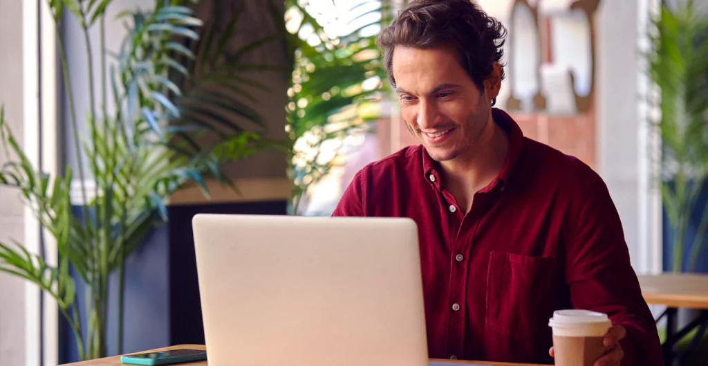 Um homem usando uma camisa vermelha, sorrindo enquanto usa um laptop e segura uma xícara de café em uma sala iluminada com plantas.