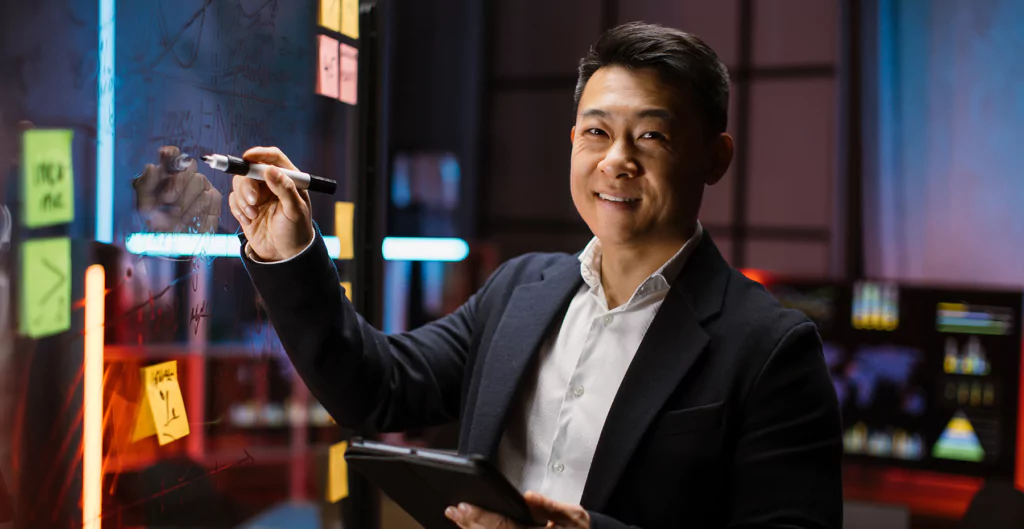 Homem asiático sorrindo enquanto escreve em um quadro transparente em ambiente moderno.